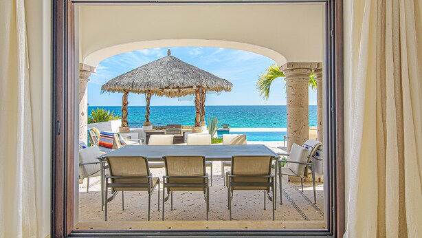 Une terrasse avec une table et des chaises, un parasol en chaume et une vue sur l’océan.