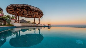 Une piscine à débordement paisible, dotée d’un parasol en chaume et d’un foyer, où se reflète le coucher de soleil sur l’océan.