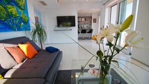 A modern living room with a grey sofa, colorful cushions, a glass coffee table, a large painting, a bookshelf, and a TV mounted on the wall. - Villa Evergreen with Pool (Crikvenica)