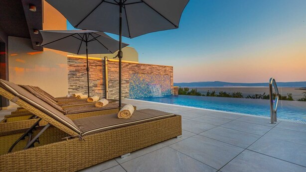 A rooftop infinity pool with lounge chairs and umbrellas, a stone wall with a mosaic, and a view of the sea at sunset.