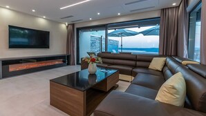 A modern living room with a large sectional sofa, a flat-screen TV, a fireplace, and a large window offering a view of the sea.