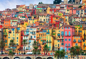 Une colline densément bâtie, parsemée de bâtiments aux couleurs vives et dotés de balcons, avec un pont de pierre au premier plan et des palmiers.