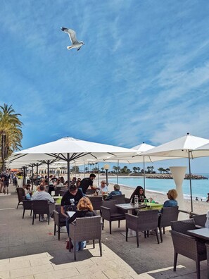 Un café en plein air, avec des tables et des parasols, offrant une vue sur une plage et la mer.