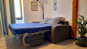 Un lit compact avec un matelas bleu et des oreillers à motifs, une table de chevet, une plante en pot et un cadre photo fixé au mur.