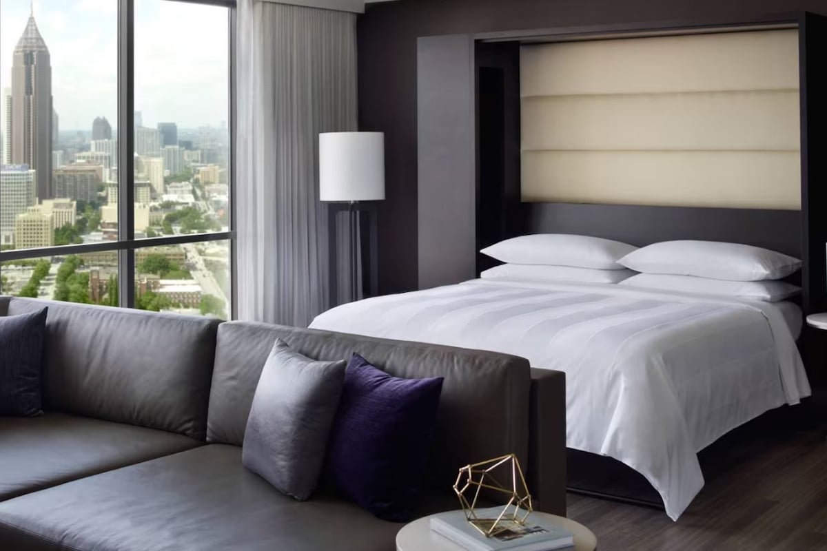 A modern hotel room with a large bed, a sofa, a city view, and a lamp.