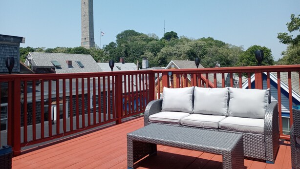 Ein Balkon mit einer Rattan-Couch und roter Geländer, mit Blick auf eine Stadt, die von einem markanten Turm und Bäumen geprägt ist.