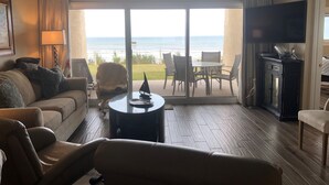 Een woonkamer met een bank, fauteuils, een salontafel en een televisie. Via grote glazen deuren is er uitzicht op de oceaan.