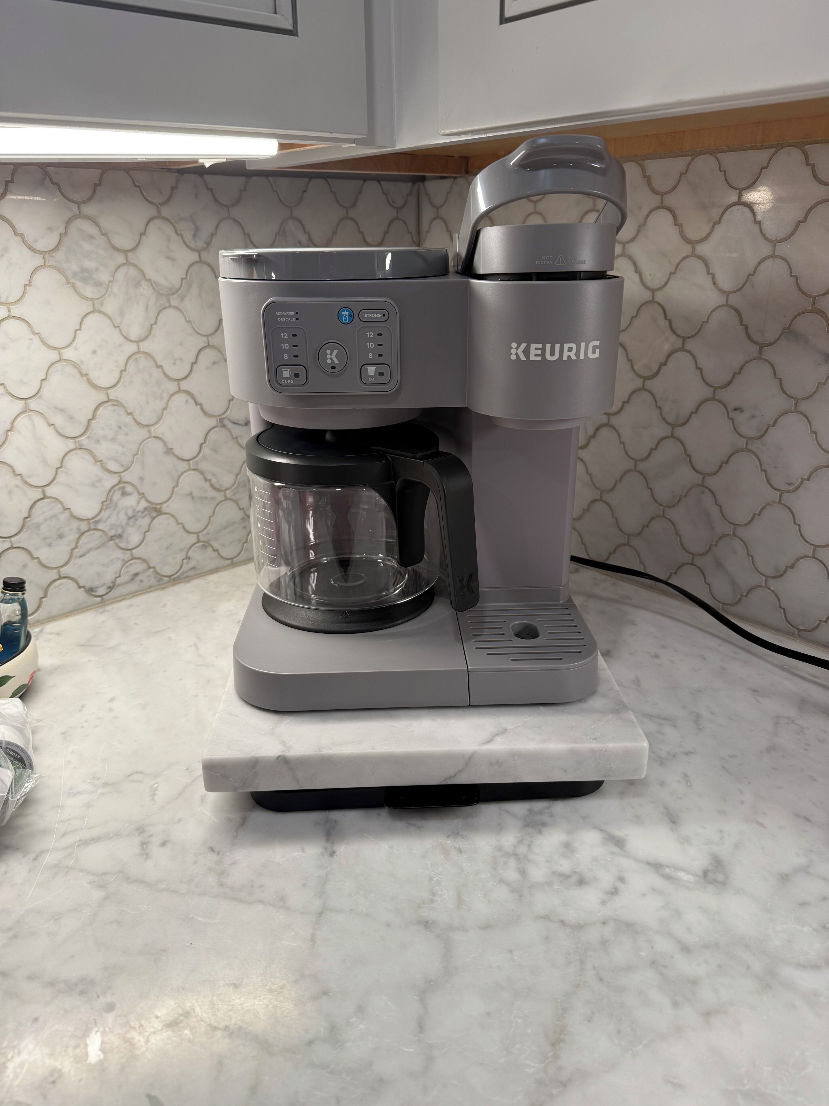Une machine à café Keurig posée sur un comptoir en marbre, avec un pare-éclaboussures à motifs.