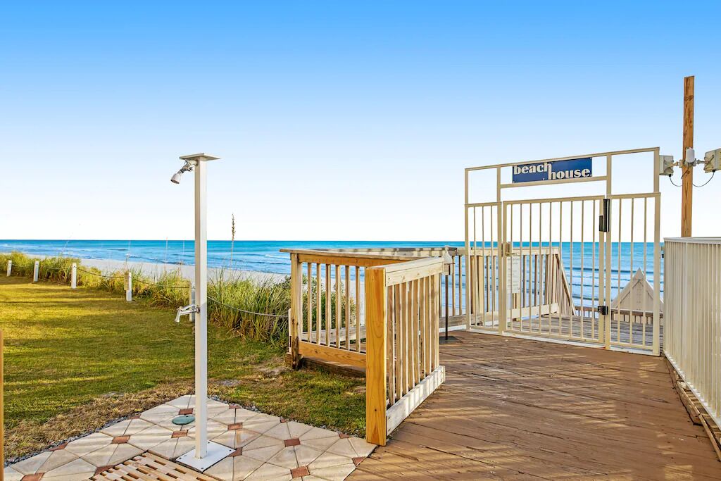 En træterrasse der fører til en låge med påskriften 'Beach House', med udsigt til havet.