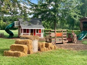 Explore a charming play area with a wooden structure, wagon, and hay bales, perfect for kids to play and climb.