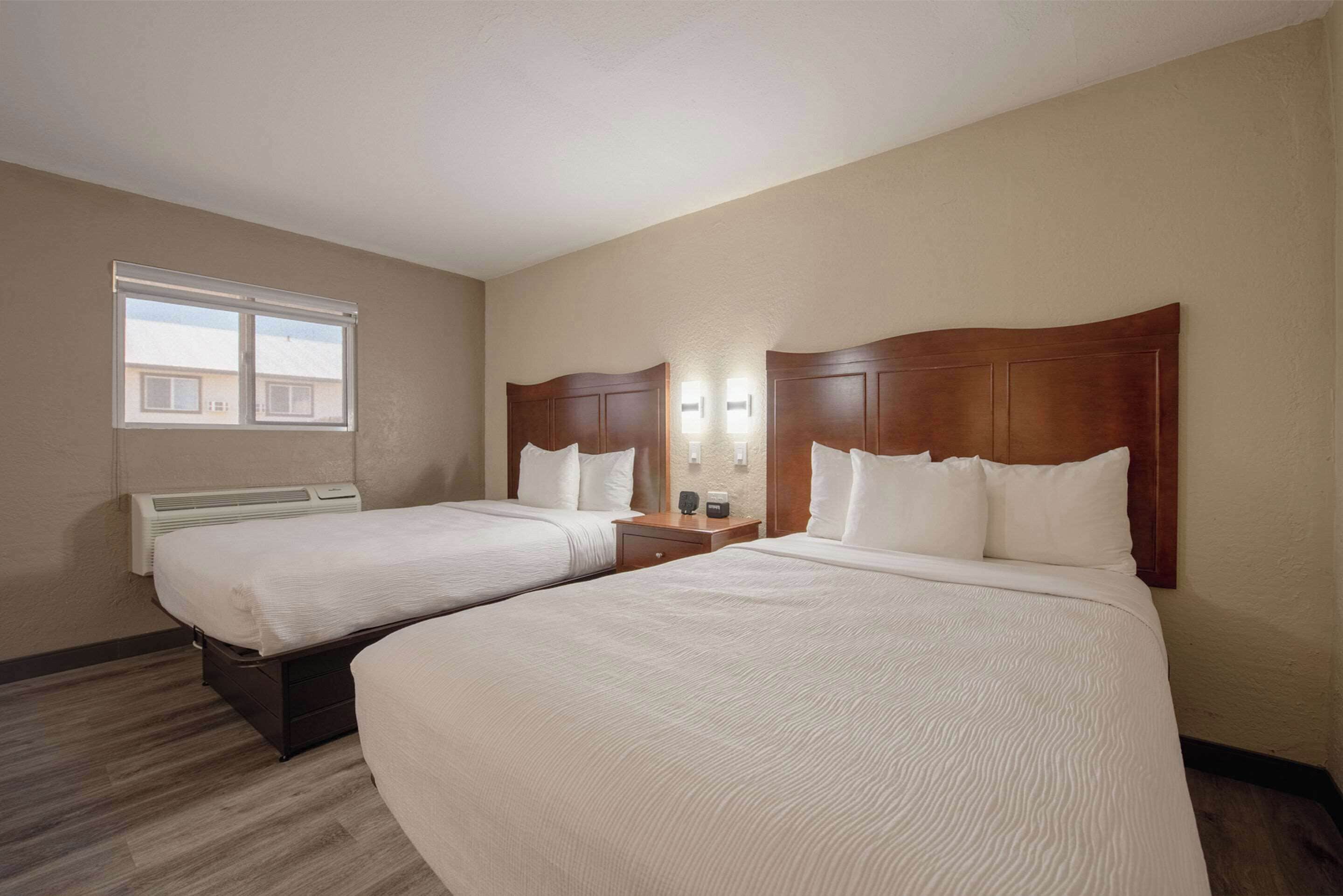 A hotel room with two beds, a window, an air conditioning unit, and a nightstand with a lamp.