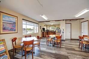 A spacious dining area with multiple wooden tables and chairs, a kitchen in the background, and large windows providing natural light.