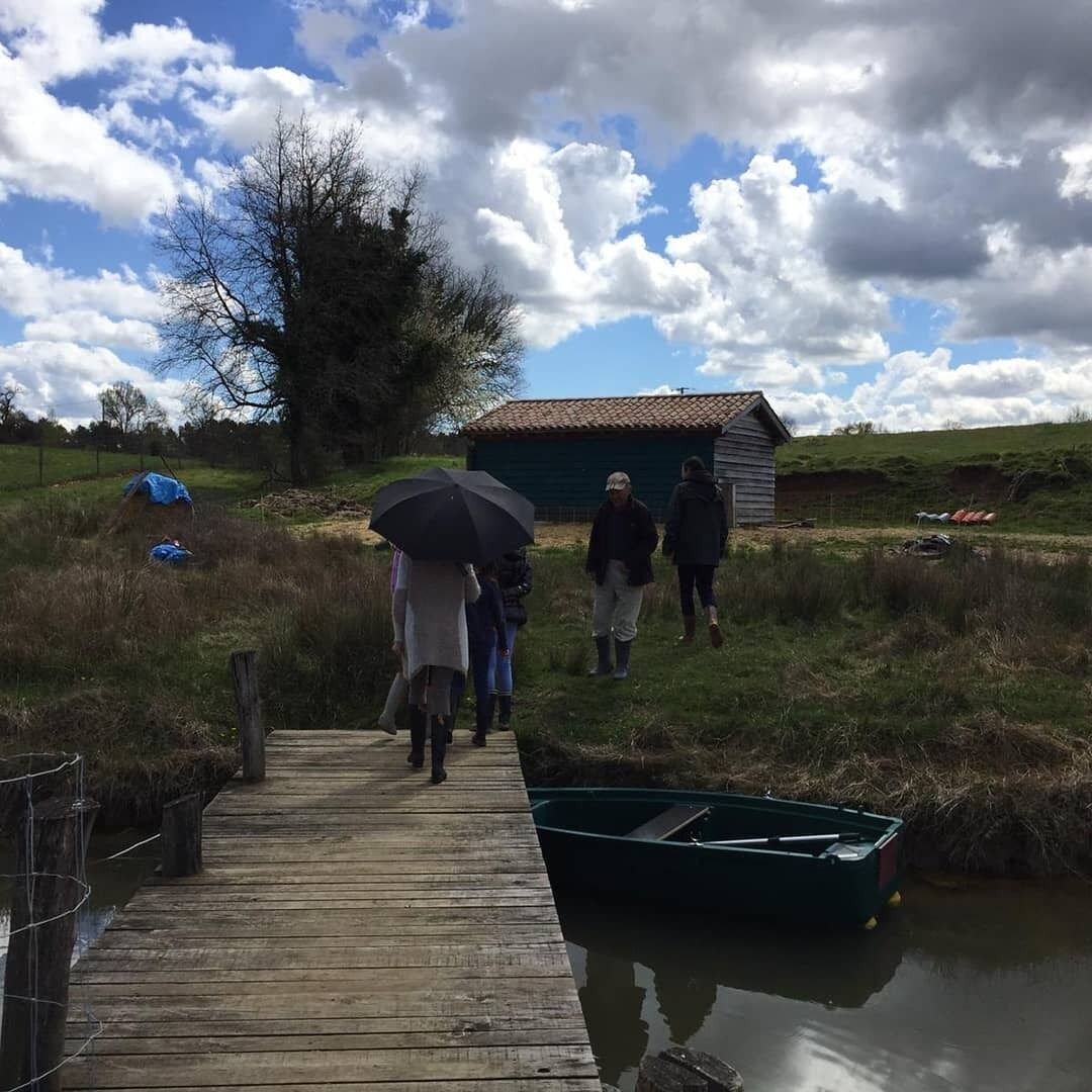 Une jetée en bois menant à un petit bateau, avec des personnes qui marchent en direction d’un bâtiment situé au loin.