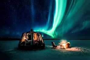A geodesic dome structure with a warm interior light, a chair, and a small fire on a snowy landscape under a starry night sky with the Northern Lights. - Peace & Quiet Hotel (Jokkmokk)