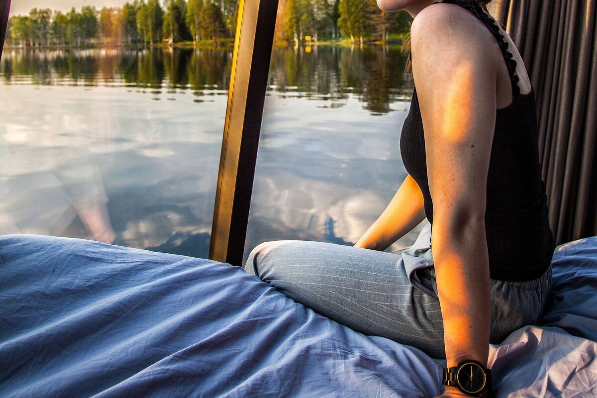 Une personne assise sur un lit, avec vue sur un lac et des arbres, à travers une fenêtre.