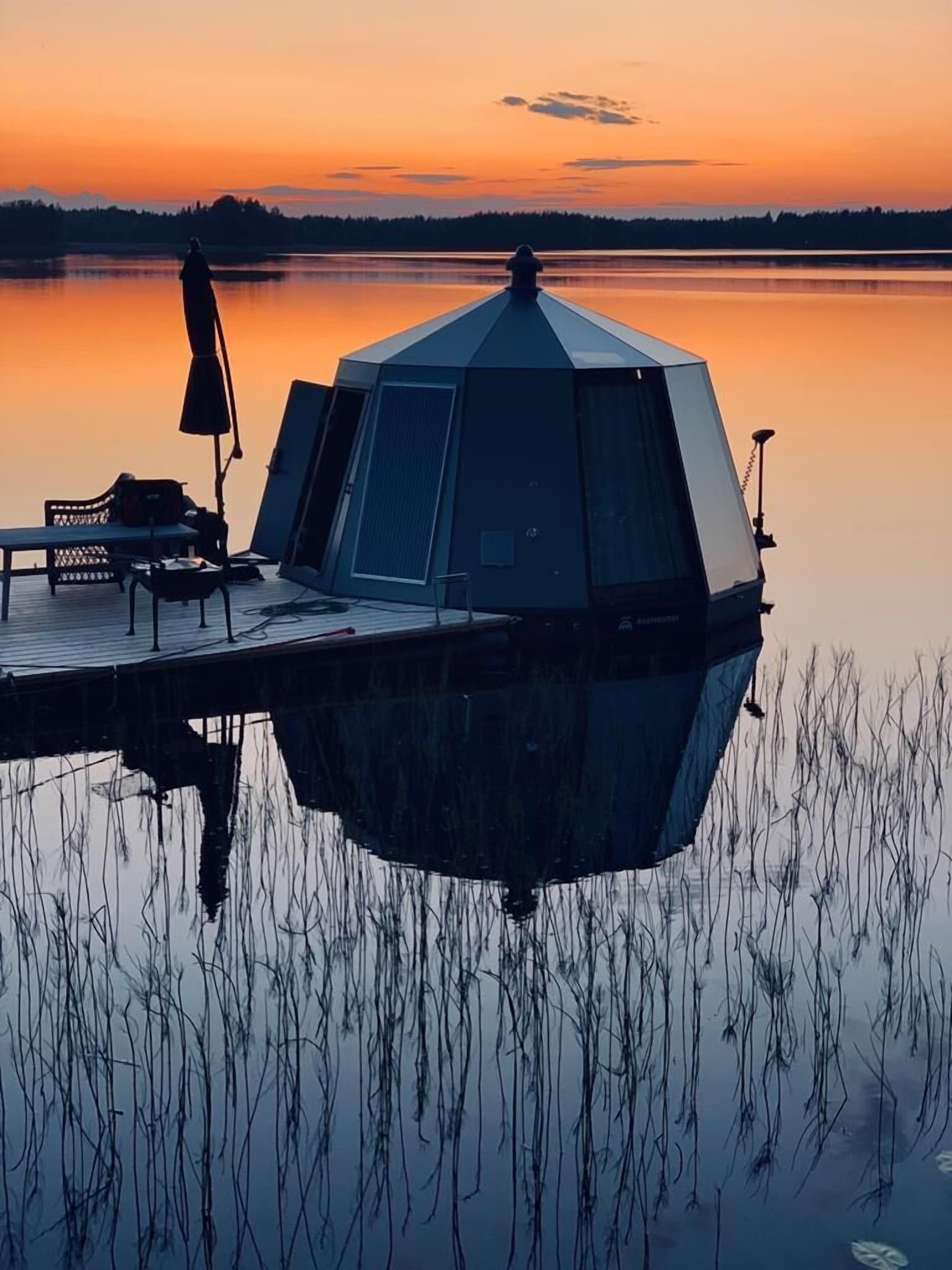 A hexagonal gazebo on a dock with a bench and umbrella, overlooking a calm lake at sunset.