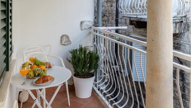 Un balcón con mesa y sillas blancas, una planta en maceta y un muro de piedra.