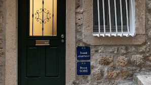 Un bâtiment en pierre avec une porte noire et une fenêtre grillagée. On y voit des panneaux indiquant un appartement et une pièce réservée à un scribe.
