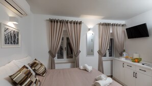 Une chambre d’hôtel avec un lit, des rideaux, un téléviseur à écran plat, une fenêtre, un tableau accroché au mur et une petite table sur laquelle se trouve un saladier rempli de fruits et quelques verres.