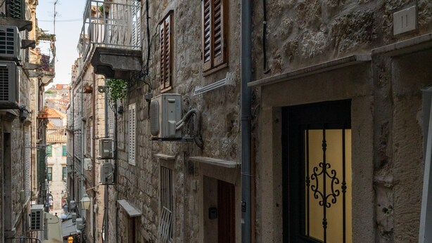 Une ruelle pavée bordée de bâtiments des deux côtés, une antenne parabolique et une porte ornée d’un cadre métallique décoratif.