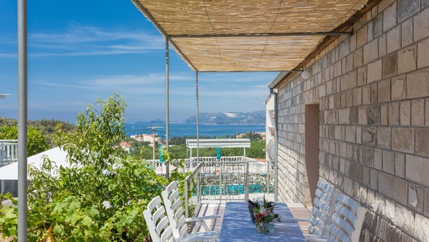 Una terrazza con tavolo e sedie, affacciata su una piscina e una vista panoramica del mare e delle montagne.