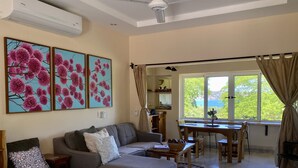 Un salon comprenant un canapé, un pouf et un coin repas avec une table et des chaises. De grandes fenêtres sont présentes, avec des rideaux, ainsi qu'un ventilateur de plafond et des œuvres d'art accrochées au mur.