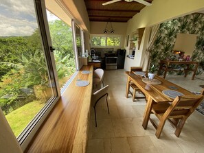 Un coin repas avec une table et des chaises en bois, une cuisine en arrière-plan et une vue sur une végétation luxuriante à l’extérieur.