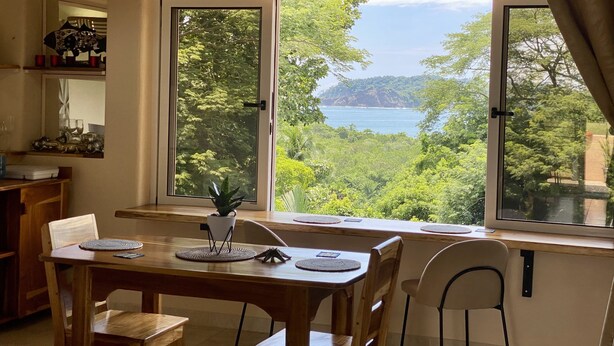 Un coin repas avec une table et des chaises, une plante en pot et une vue sur un paysage côtier, visible à travers de grandes fenêtres.