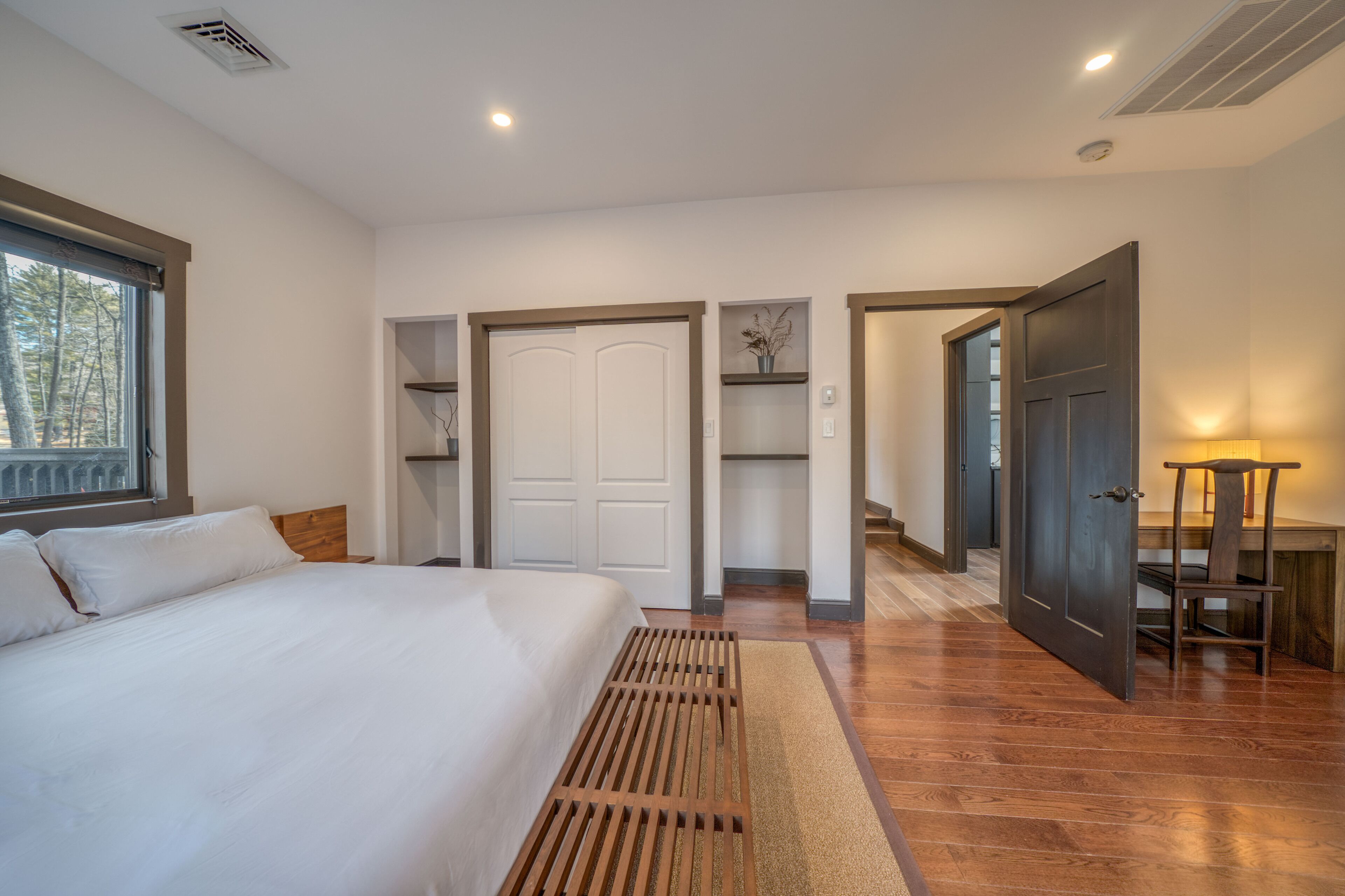 Un dormitorio con una cama grande, cabecera de madera, una ventana con persianas, una puerta que da a un pasillo y una pequeña mesa con una silla.
