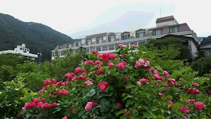 A building with multiple windows and a flat roof, surrounded by lush greenery and pink flowers. - Hotel Koryu (Fujikawaguchiko)