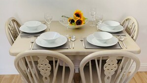 Una mesa de comedor dispuesta con platos, tazones y cubiertos blancos, rodeada de sillas blancas con detalles decorativos.