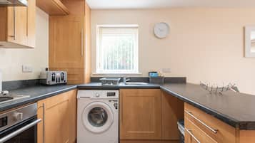 A compact kitchen with wooden cabinets, a built-in washing machine, a toaster, and a window with blinds.