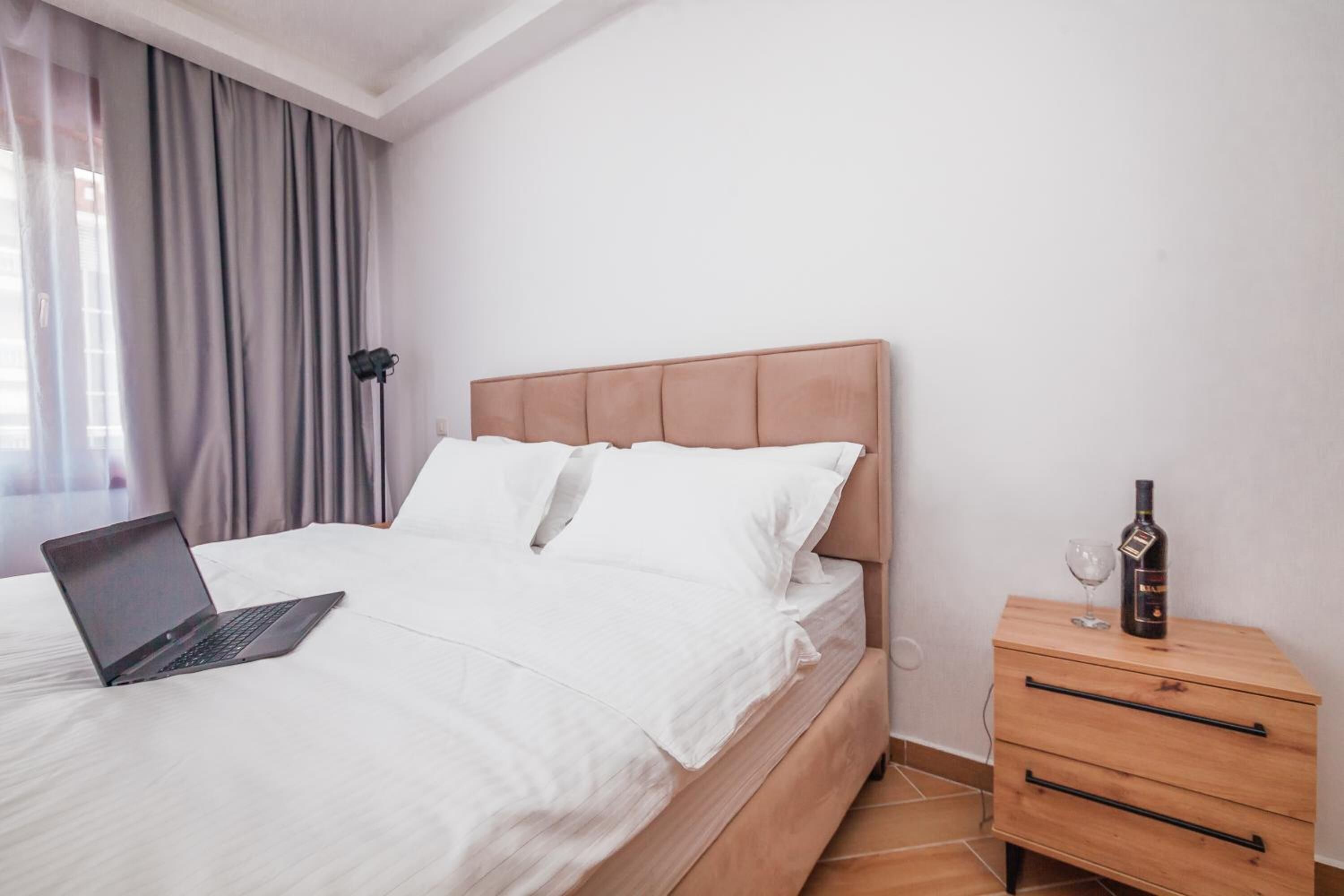 A bedroom with a bed, a laptop on the bed, a bedside table with a bottle and glass, and a lamp.