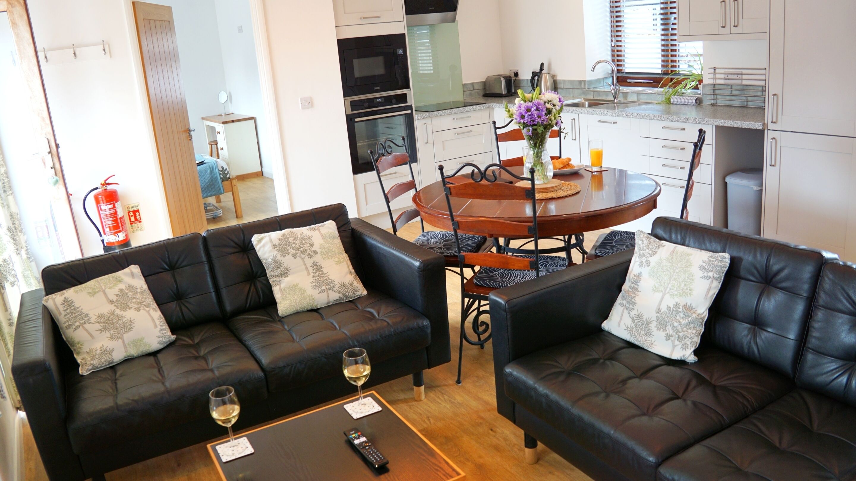 A living room with two black leather sofas, a wooden dining table with chairs, and a kitchen in the background.