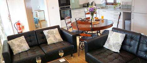 A living room with two black leather sofas, a wooden dining table with chairs, and a kitchen in the background.