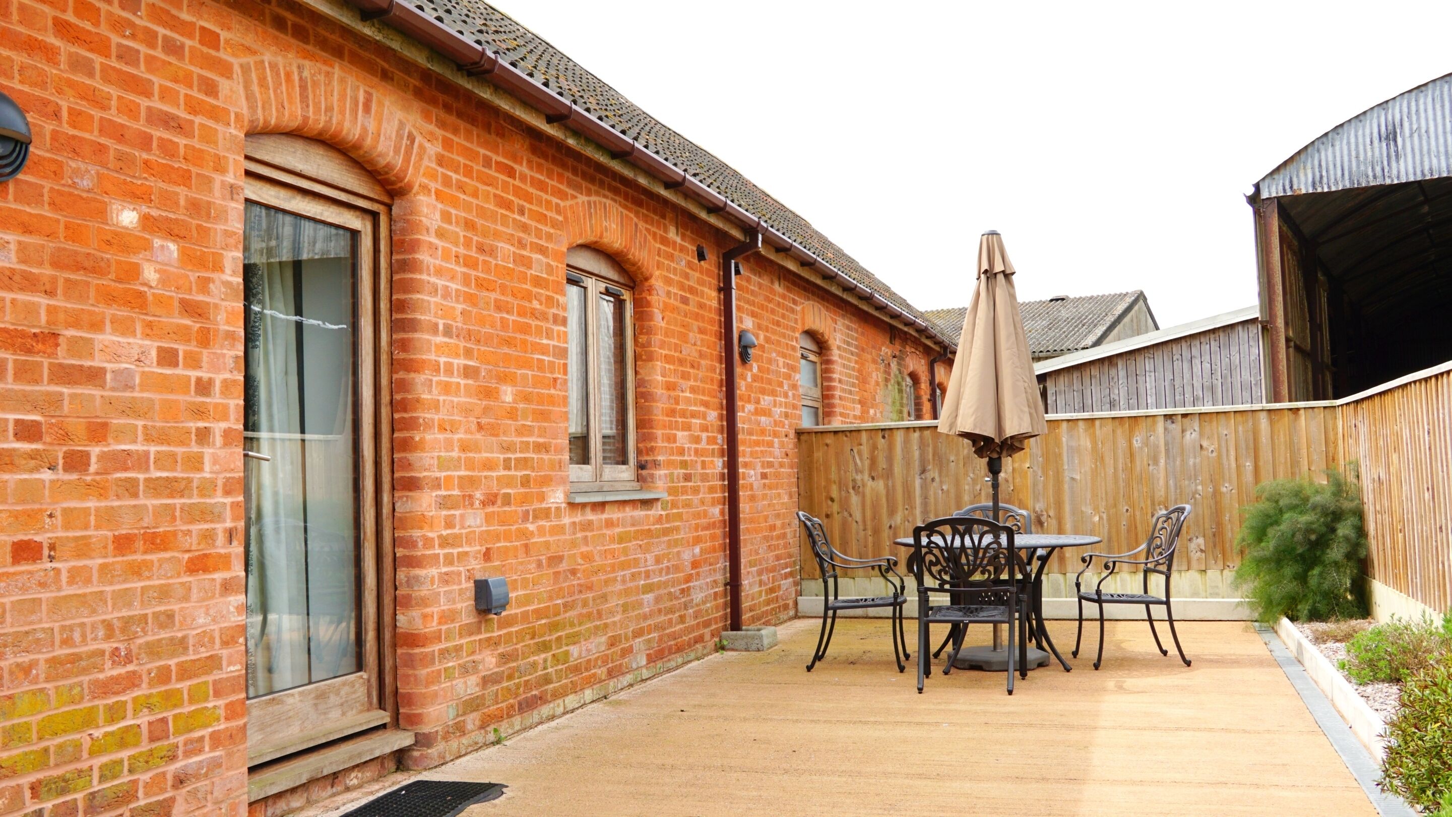 A brick building with a patio area featuring a table and chairs, a wooden fence, and a covered umbrella.