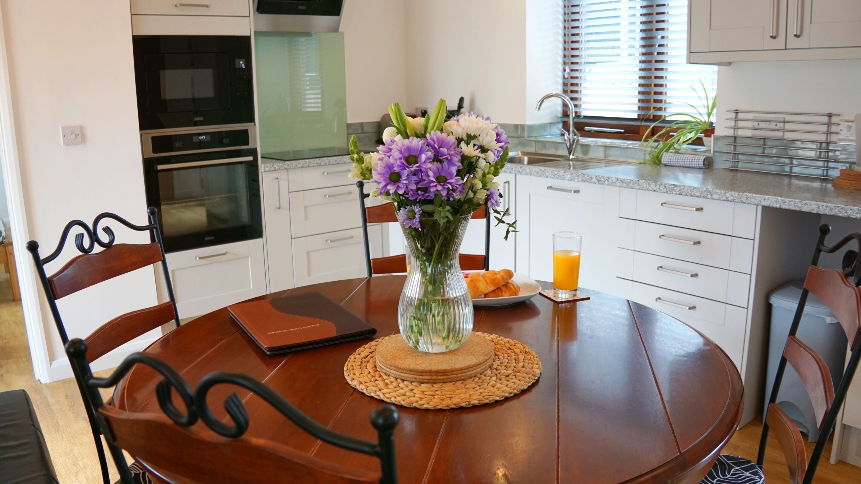 A dining area with a round wooden table, chairs, a vase with flowers, a glass of orange juice, and a plate with food.