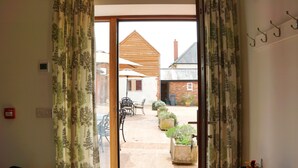 A room with a sliding glass door leading to an outdoor patio area with a wooden table and chairs, potted plants, and a brick building in the background. - Courtbrook Farm Apartments (Exeter)