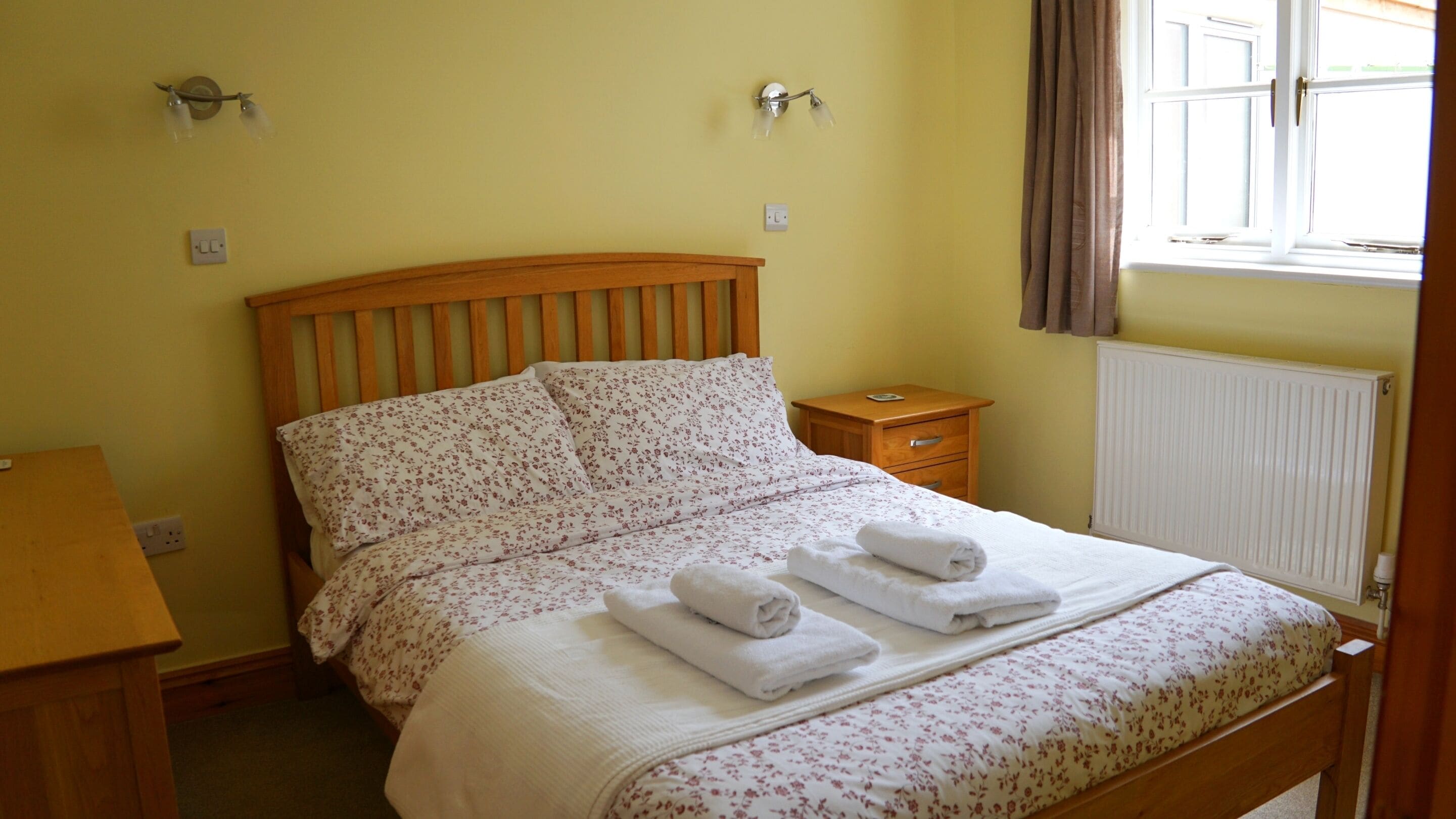 Une chambre à coucher avec un lit en bois, une table de chevet, une commode, un radiateur et une fenêtre avec des rideaux.