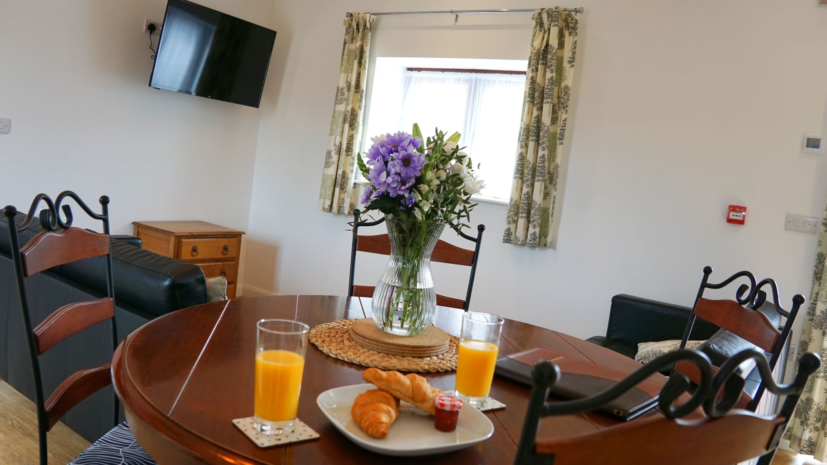 A dining table with chairs, a vase with flowers, a plate with croissants and jam, and glasses of orange juice.