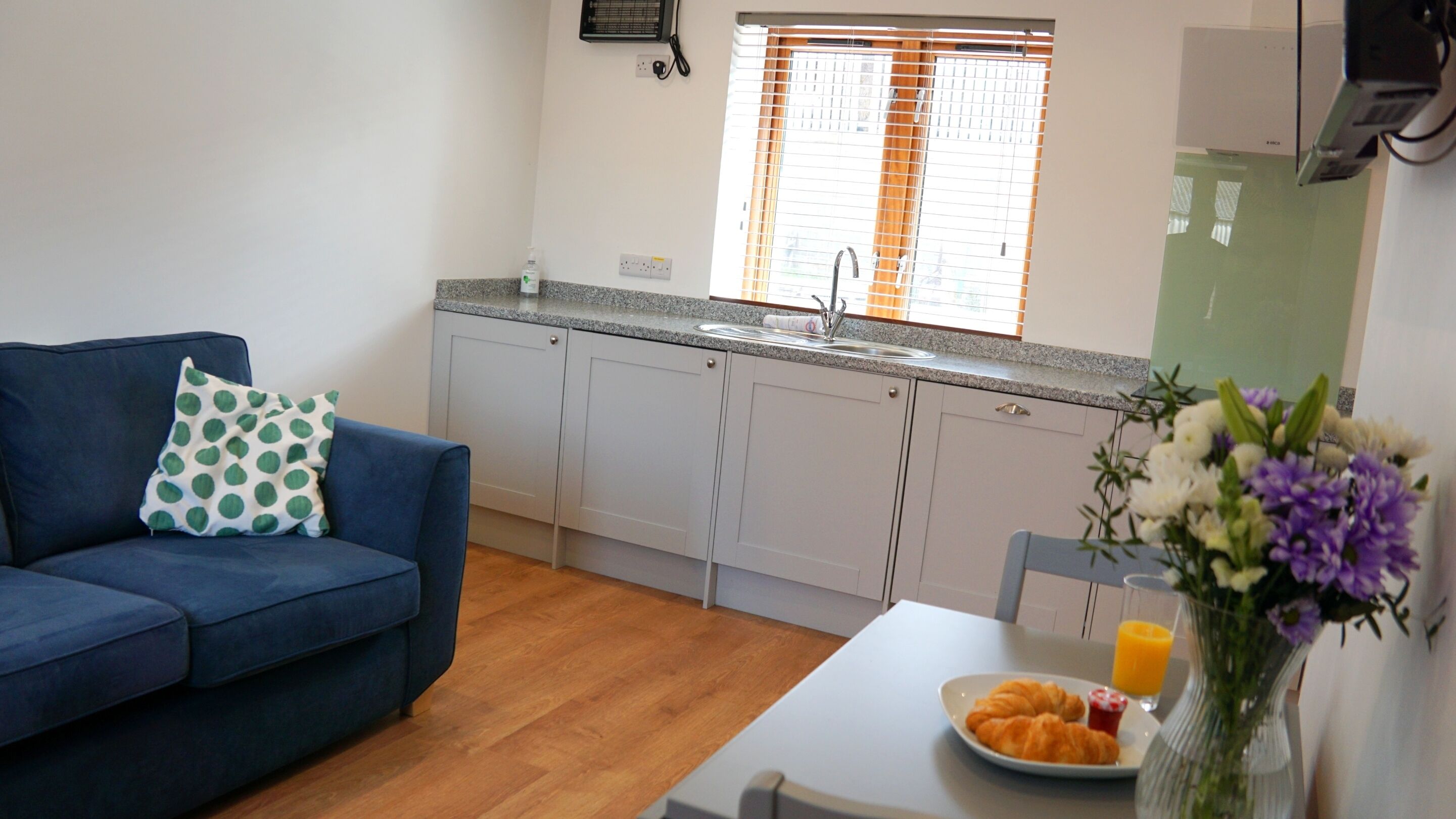 A compact living space with a kitchenette, a sofa with a polka dot pillow, a dining table with croissants and juice, and a vase with flowers.