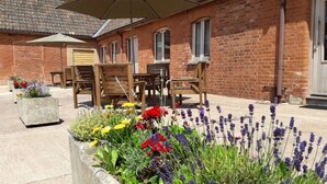 A brick building with a patio area featuring outdoor furniture and flower beds. - Courtbrook Farm Apartments (Exeter)