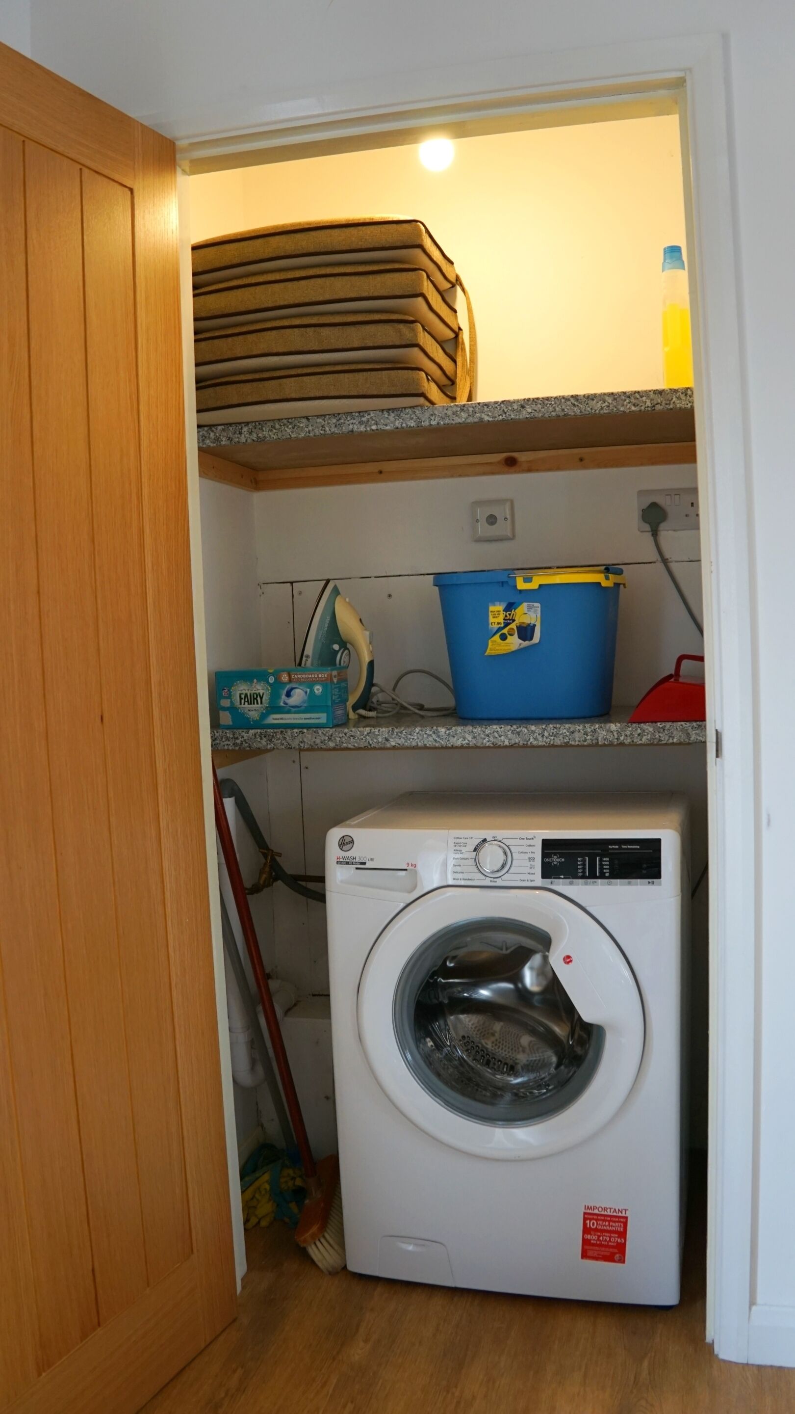 Uma área de lavanderia com máquina de lavar, um balde azul e uma caixa de detergente Fairy.