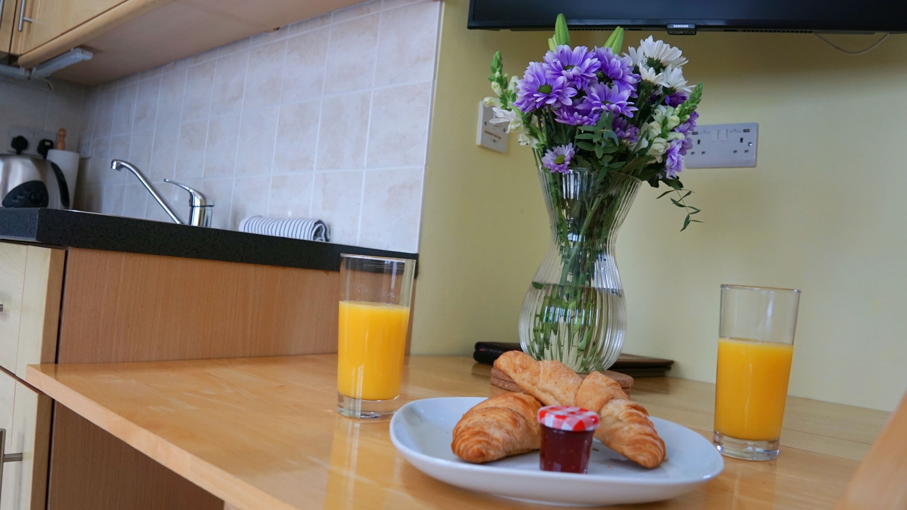 Ein Frühstückstisch mit Croissants, Marmelade und Orangensaft, eine Blumenvase und eine Küchenarbeitsplatte.
