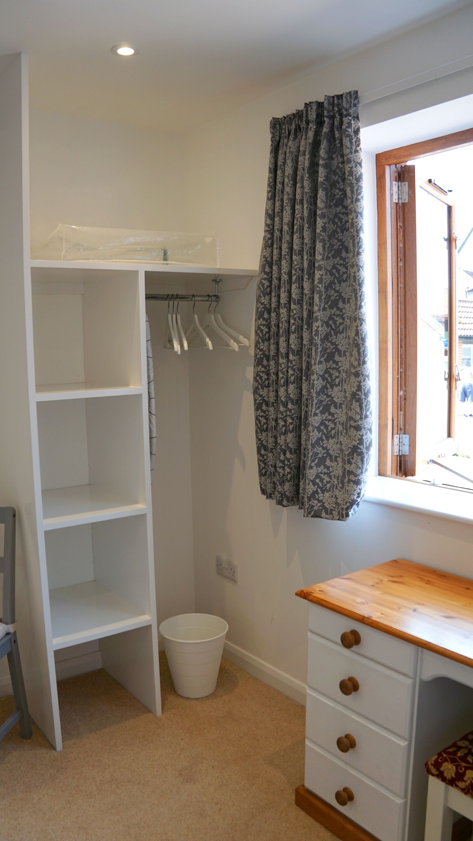 A room with a white wardrobe, a wooden desk, a hanging coat rack, and a window with curtains.