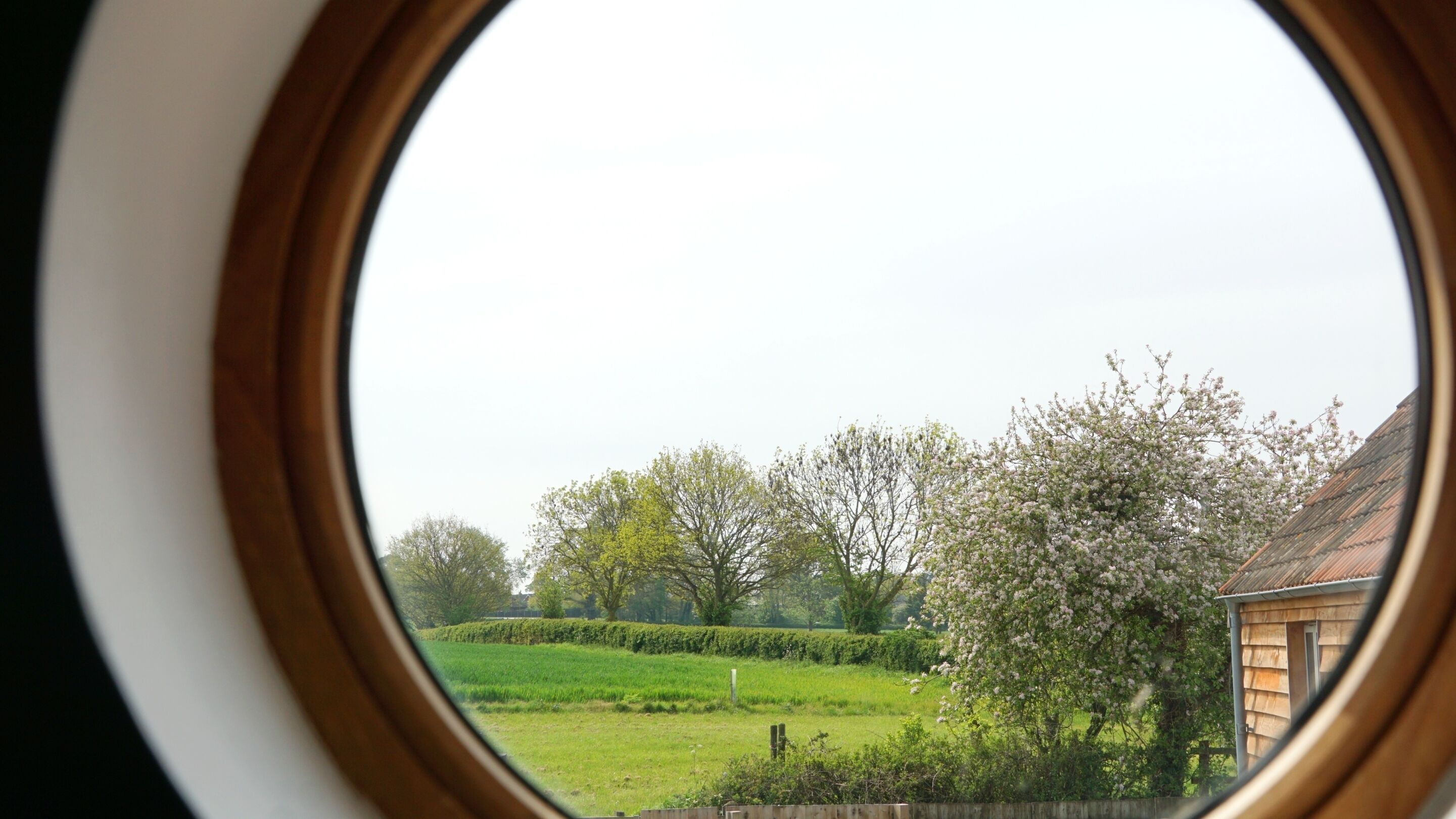 Blick durch ein rundes Fenster auf ein grünes Feld, Bäume und ein Holthaus.