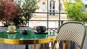 A round table with two cups and saucers, a wicker chair, and a building with arched windows in the background. - Bonsoir Madame (Paris)