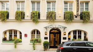 La façade d’un hôtel, ornée d’arcs en plein cintre au niveau des fenêtres, avec une entrée centrale et des plantes vertes sur les balcons.