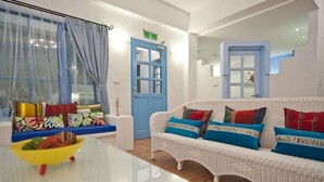 Una habitación con un sofá de mimbre blanco, cojines coloridos, una mesa de vidrio con un tazón amarillo y una puerta azul.