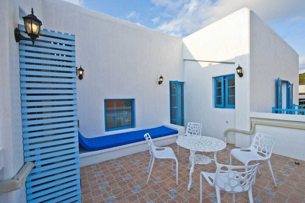 Un edificio blanco con contraventanas azules y una pequeña zona de estar con sillas y una mesa blancas.